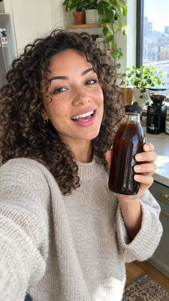 A smiling woman with curly hair in a sunny kitchen holds an amber bottle of cold brew toward her phone camera